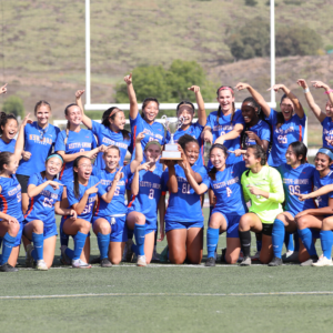 The Pomona-Pitzer women's soccer team gather with their coaches on the field in two rows while raising their hands triumphantly. 中间的一名球员举着奖杯. The team wears blue uniforms with white and orange accents.
